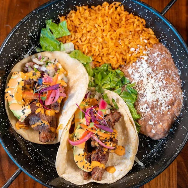 Two carne asada tacos with rice, refried beans, and lettuce on a black plate.